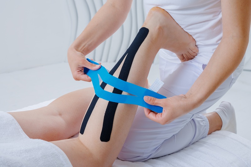 Closeup of therapist applying kinesiology tape on patient’s leg to reduce strain, improve mobility, and support recovery from muscle injury.